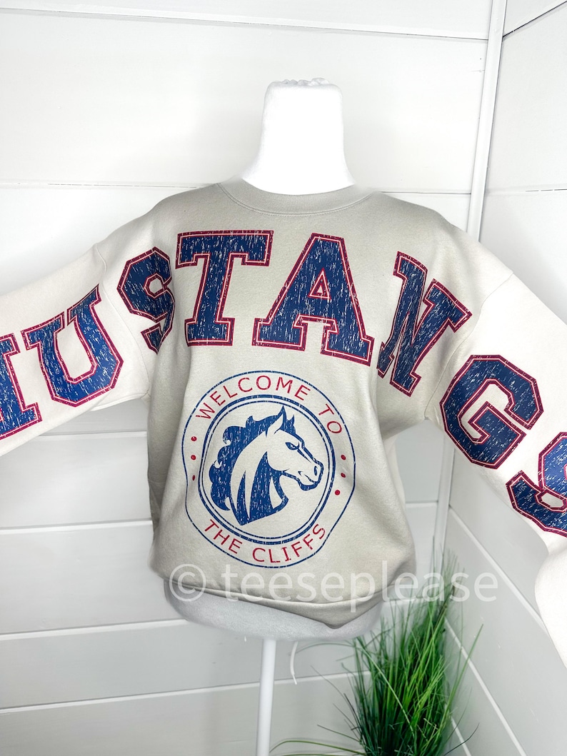 May include: A beige crewneck sweatshirt featuring the word MUSTANGS in large, distressed navy blue letters with red outlines. A circular graphic on the chest displays a horse head profile with the text WELCOME TO THE CLIFFS in navy blue.