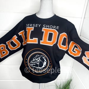 May include: A black long-sleeve sweatshirt featuring the text JERSEY SHORE in white and the word BULLDOGS in large orange letters with white outlines. A circular graphic below displays a white bulldog head illustration on a black background.