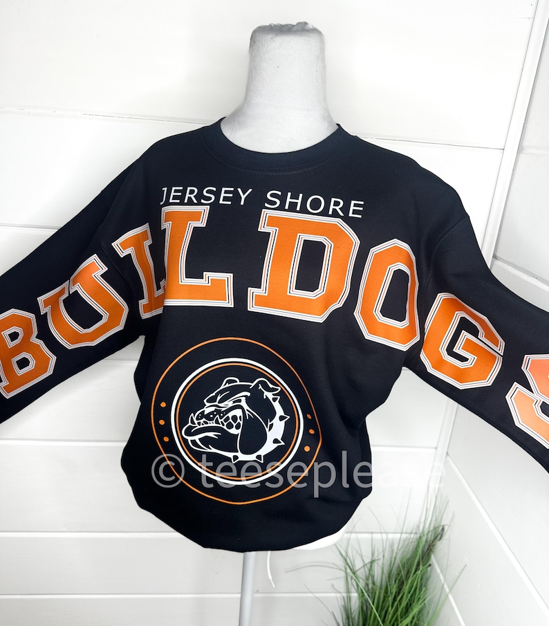 May include: A black long-sleeve sweatshirt featuring the text JERSEY SHORE in white and the word BULLDOGS in large orange letters with white outlines. A circular graphic below displays a white bulldog head illustration on a black background.