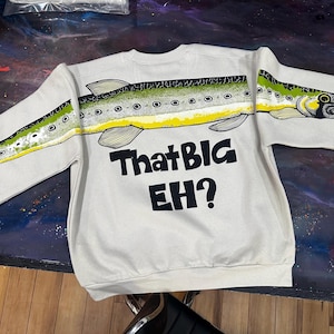 May include: A cream-colored crewneck sweatshirt featuring a continuous graphic of a green and yellow fish spanning across the chest and sleeves. Below the fish, the text "That BIG EH?" is printed in bold black lettering on the front of the garment.