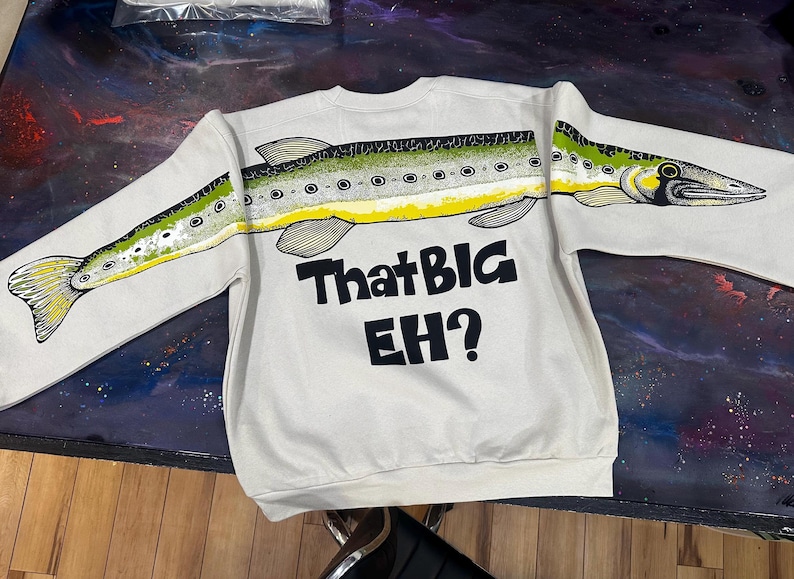 May include: A cream-colored crewneck sweatshirt featuring a continuous graphic of a green and yellow fish spanning across the chest and sleeves. Below the fish, the text "That BIG EH?" is printed in bold black lettering on the front of the garment.