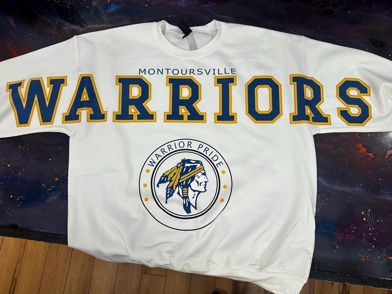 May include: A white long-sleeve sweatshirt featuring the text MONTOURSVILLE WARRIORS in large blue letters with yellow outlines. Below the text is a circular logo containing the words WARRIOR PRIDE and an illustration of a person in a headdress.