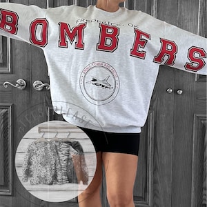 May include: A light gray crewneck sweatshirt featuring the red text BOMBERS across the chest and sleeves. Above the text is FREDERICK, OK. Below is a circular emblem with a jet illustration and the text SMALL TOWN BIG PRIDE.