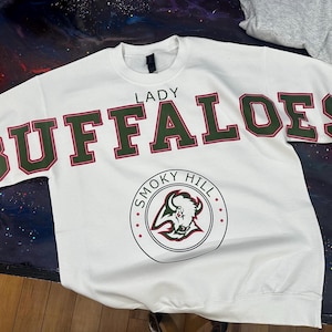 May include: A white long-sleeve sweatshirt featuring the text LADY BUFFALOES in large green letters with red outlines across the chest. Below the text is a circular logo containing the words SMOKY HILL and a graphic of a buffalo head in red and green.