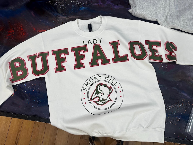 Op de afbeelding: Witte sweatshirt met de tekst "LADY BUFFALOES" in grote, groen en rode letters. Onder de tekst staat een cirkelvormig logo met een buffelkop en de woorden "SMOKY HILL".