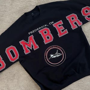 Op de afbeelding: Zwart sweatshirt met het woord "BOMBERS" in rode blokletters met witte rand op de mouwen en over de borst. De tekst "Frederick, OK" staat boven een cirkelvormige afbeelding met een vliegtuig en de woorden "SMALL TOWN BIG PRIDE".