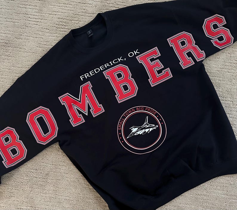 Op de afbeelding: Zwart sweatshirt met het woord "BOMBERS" in rode blokletters met witte rand op de mouwen en over de borst. De tekst "Frederick, OK" staat boven een cirkelvormige afbeelding met een vliegtuig en de woorden "SMALL TOWN BIG PRIDE".