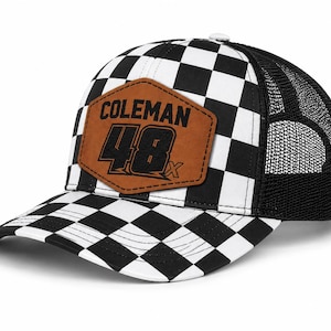 May include: A black and white chequered baseball cap with a mesh back. The cap has a brown leather patch with the name "Coleman" and the number "48x" in black lettering. The peak is also chequered.