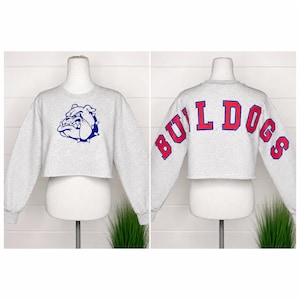 May include: A light gray cropped sweatshirt featuring a blue bulldog graphic on the front and the word BULLDOGS in red letters with blue outlines arched across the back. The garment has long sleeves and a raw-cut hemline at the waist.
