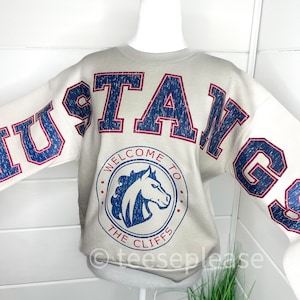 Op de afbeelding: Een lichtgrijs sweatshirt met het woord "MUSTANGS" in blauwe letters met rode rand over de borst en mouwen. Een cirkelvormige afbeelding in het midden leest "WELCOME TO THE CLIFFS" rond een blauwe paardenkop.
