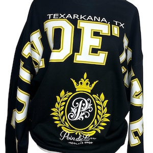 May include: A black long-sleeve sweatshirt featuring the text TEXARKANA, TX and large white block letters. The center displays a yellow crown above a circular emblem with a stylized letter P, surrounded by leaves, and the text Poin de Luxe CHOCOLATE SHOP.