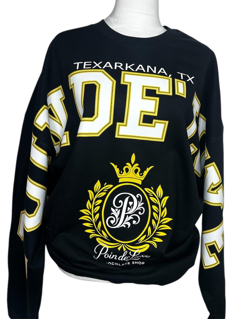 May include: A black long-sleeve sweatshirt featuring the text TEXARKANA, TX and large white block letters. The center displays a yellow crown above a circular emblem with a stylized letter P, surrounded by leaves, and the text Poin de Luxe CHOCOLATE SHOP.