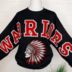 May include: A black long-sleeve sweatshirt featuring the word WARRIORS in large red block letters with white outlines. Below the text is a graphic illustration of a person wearing a traditional feathered headdress in red, white, and black.