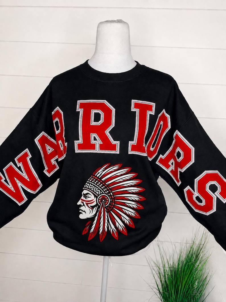 May include: A black long-sleeve sweatshirt featuring the word WARRIORS in large red block letters with white outlines. Below the text is a graphic illustration of a person wearing a traditional feathered headdress in red, white, and black.