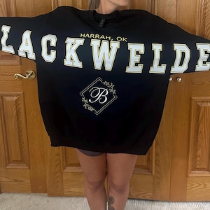 May include: A black crewneck sweatshirt featuring the text BLACKWELDER in large white block letters with gold outlines across the chest. Above the text is HARRAH, OK in gold. Below is a gold diamond frame containing a white letter B with floral accents.