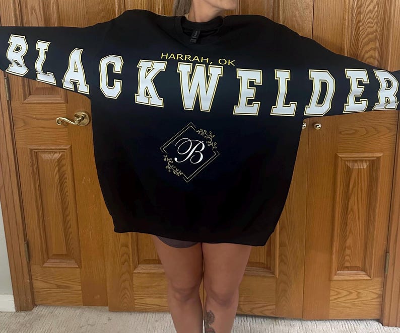May include: A black crewneck sweatshirt featuring the text BLACKWELDER in large white block letters with gold outlines across the chest. Above the text is HARRAH, OK in gold. Below is a gold diamond frame containing a white letter B with floral accents.