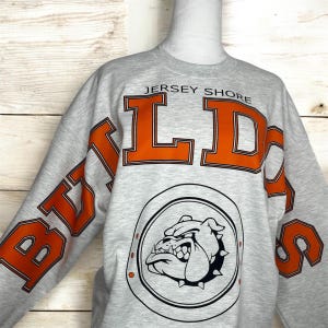 May include: A light gray crewneck sweatshirt featuring the text JERSEY SHORE in small black letters above large orange block letters that spell BULLDOGS. Below the text is a circular graphic containing a black and white illustration of a bulldog.