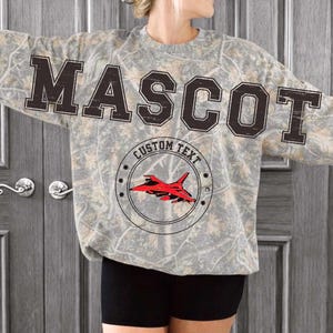 May include: A long-sleeve sweatshirt with a camouflage pattern in shades of gray, brown, and tan. The word MASCOT is printed in large black block letters across the chest. Below, a circular emblem features a red fighter jet and the text CUSTOM TEXT.