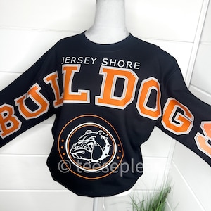 Op de afbeelding: Zwart sweatshirt met oranje en witte tekst en afbeeldingen. De tekst luidt "Jersey Shore Bulldogs" met een bulldogafbeelding in een cirkel. De mouwen hebben ook het woord "Bulldogs" in oranje en wit. Het sweatshirt wordt op een mannequin getoond.
