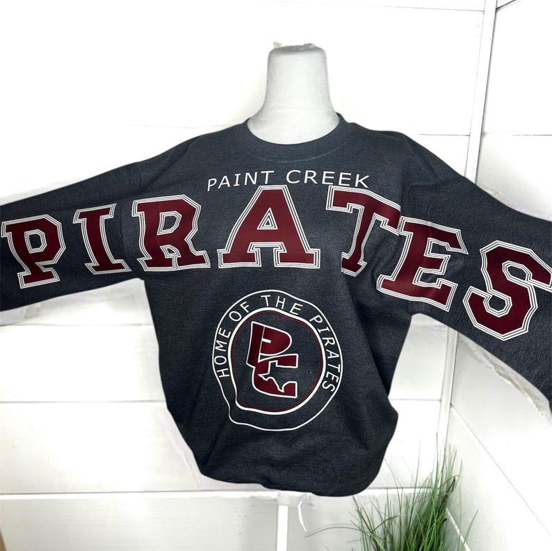 May include: A dark gray long-sleeve sweatshirt featuring the text PAINT CREEK PIRATES in large maroon letters with white outlines. Below the text is a circular logo containing the letters PC and the words HOME OF THE PIRATES in white.