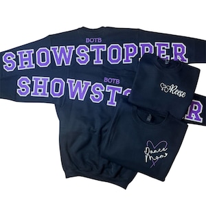 Op de afbeelding: Verschillende zwarte sweatshirts met witte en paarse letters. E&eacute;n sweatshirt heeft de tekst "SHOWSTOPPER" en "SHOWST" in grote letters. Andere sweatshirts hebben de namen "Reese" en "Dance Mom" in cursief.