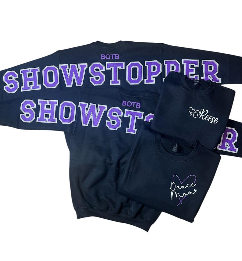 Op de afbeelding: Verschillende zwarte sweatshirts met witte en paarse letters. E&eacute;n sweatshirt heeft de tekst "SHOWSTOPPER" en "SHOWST" in grote letters. Andere sweatshirts hebben de namen "Reese" en "Dance Mom" in cursief.