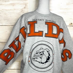 May include: A light gray crewneck sweatshirt featuring the text JERSEY SHORE in small black letters above large orange block letters spelling BULLDOGS. Below the text is a circular emblem containing a black and white illustration of a bulldog.