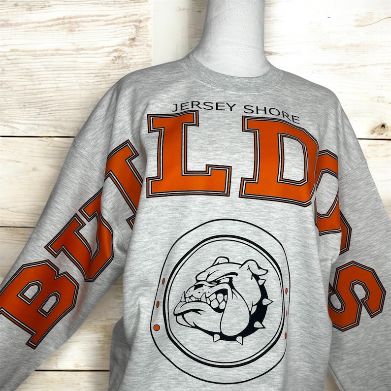 May include: A light gray crewneck sweatshirt featuring the text JERSEY SHORE in small black letters above large orange block letters spelling BULLDOGS. Below the text is a circular emblem containing a black and white illustration of a bulldog.