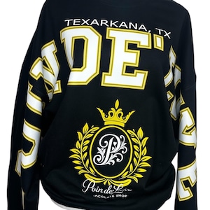 May include: A black long-sleeve sweatshirt featuring the white text TEXARKANA, TX above large white and yellow block letters. A central yellow crown and laurel wreath emblem surrounds a white letter P, with the text Poin de Luxe CHOCOLATE SHOP below.