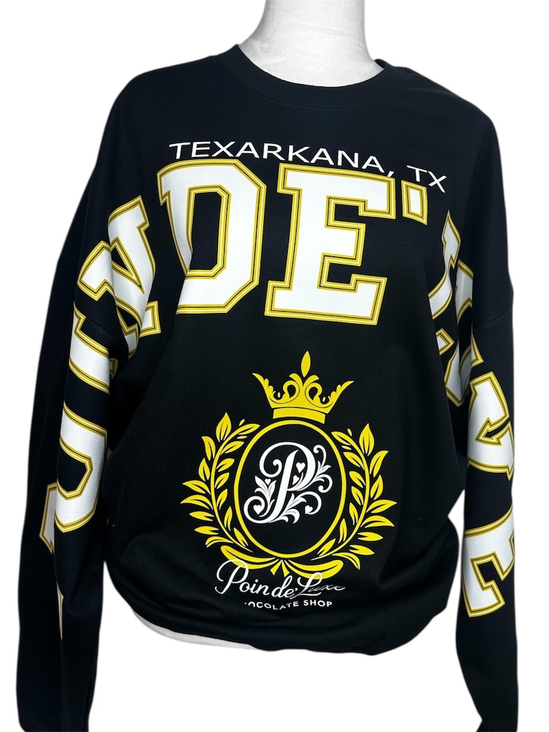 May include: A black long-sleeve sweatshirt featuring the white text TEXARKANA, TX above large white and yellow block letters. A central yellow crown and laurel wreath emblem surrounds a white letter P, with the text Poin de Luxe CHOCOLATE SHOP below.