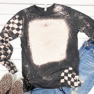 May include: A black long-sleeved shirt with a white bleach design. The design features a large square in the center and checkered patterns on the sleeves and bottom right corner.