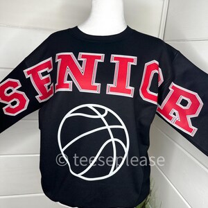 May include: A black long-sleeve sweatshirt featuring the word SENIOR in large red block letters with white outlines across the chest and sleeves. A white graphic of a basketball is centered below the text. The text teeseplease is visible.