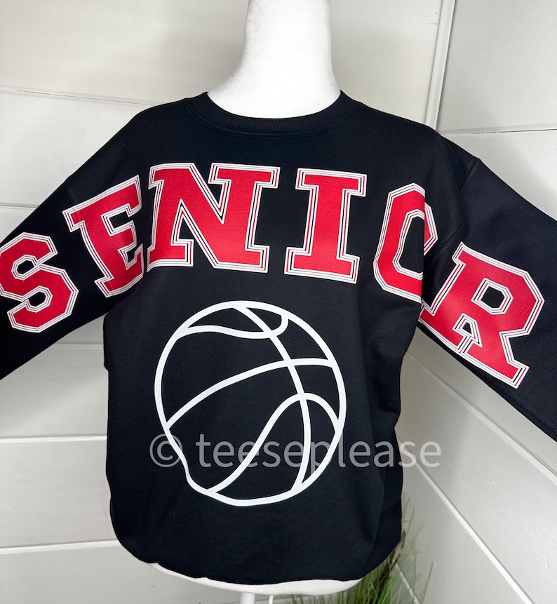 May include: A black long-sleeve sweatshirt featuring the word SENIOR in large red block letters with white outlines across the chest and sleeves. A white graphic of a basketball is centered below the text. The text teeseplease is visible.