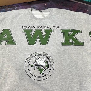 May include: A light gray crewneck sweatshirt featuring the text IOWA PARK, TX and HAWKS in large dark green block letters. Below the text is a circular emblem containing a hawk graphic over a map of Texas with the text SMALL TOWN BIG PRIDE.