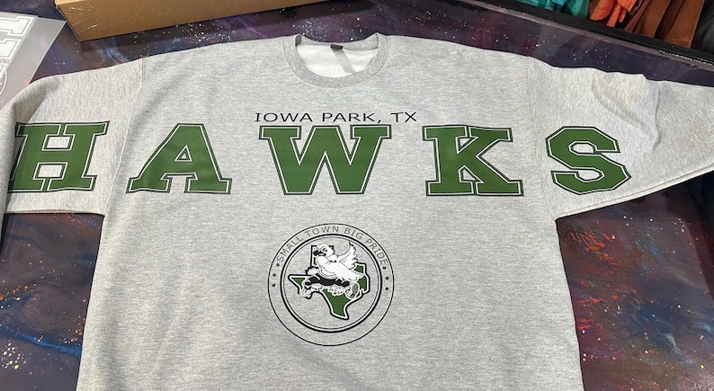May include: A light gray crewneck sweatshirt featuring the text IOWA PARK, TX and HAWKS in large dark green block letters. Below the text is a circular emblem containing a hawk graphic over a map of Texas with the text SMALL TOWN BIG PRIDE.