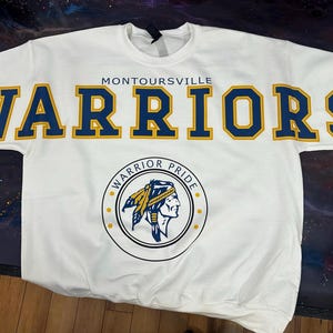 Op de afbeelding: Witte sweatshirt met het woord "WARRIORS" in grote, blauwe en gouden letters over de borst. Het woord "Montoursville" staat boven het woord "WARRIORS". Een cirkelvormige afbeelding met een Indiaans profiel staat eronder.