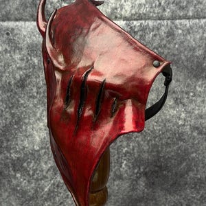 May include: A deep red, moulded mask featuring a pointed, horn-like protrusion at the top and three vertical, slash-like cutouts across the front surface. The mask has a glossy, textured finish and is secured with a black strap attached by metal rivets.