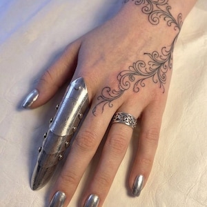 Puede incluir: Una mano con esmalte de uñas plateado y una armadura de dedo plateada. Un tatuaje de un diseño de vid en espiral está en la muñeca y la mano. Un anillo plateado con un patrón decorativo está en el dedo anular.
