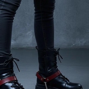 Op de afbeelding: Zwarte leren combat boots met rode details. De laarzen hebben een vetersluiting, een dikke zool en een rode band met een zilveren ketting. Gedragen met een zwarte skinny jeans.