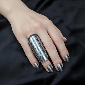May include: An articulated silver ring with an engraved floral pattern. The ring, covering the finger, is made of multiple segments and small rivets. The hand has matching silver nail polish, set against a dark background. The ring's design is intricate and detailed.