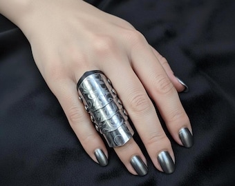 May include: A silver-toned, articulated ring with an ornate, engraved floral design. The ring covers the entire finger, featuring multiple segments and small rivet details. The ring is shown on a hand with matching silver nail polish, set against a dark background.