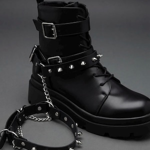 May include: A black ankle boot featuring a thick lug sole, black laces, and a silver-tone buckle strap at the ankle. A detachable black strap with silver-tone metal spikes and a silver chain connects the boot to a matching spiked ankle cuff.