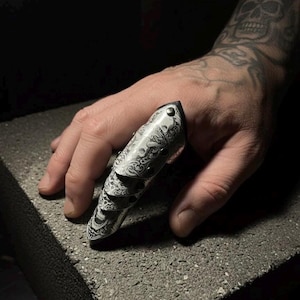 May include: An articulated finger armour ring in silver, featuring an engraved, ornate design. The ring, shown on a hand, covers the entire finger with multiple segments for flexibility. The ring is resting on a grey surface.