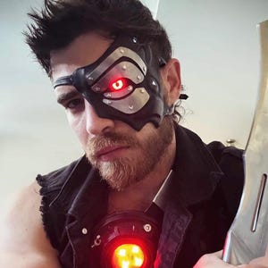 May include: A black leather eye mask with silver metal accents and a glowing red eye. The mask is worn over one eye. A red light is also visible on the chest. The image is of a person.