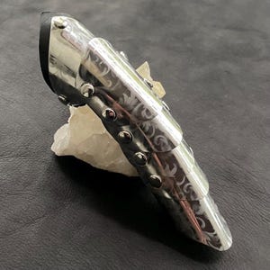 May include: A silver finger armour with a black leather base and riveted details. The metal is etched with a swirling floral pattern. The armour is designed to protect the finger, likely for cosplay or historical reenactment.