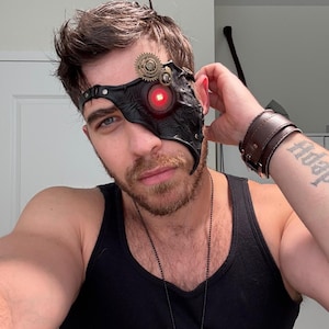 May include: A man wearing a black tank top and a steampunk-style leather eye patch with a red LED light. The eye patch has gears and other metal accents.