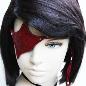 May include: A red leather eye patch with a star-shaped design and silver studs. The eye patch is worn over the left eye of a mannequin with short, dark brown hair.