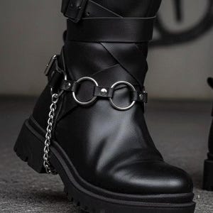 May include: Black leather boot with a chunky sole, featuring multiple straps and silver-tone metal accents. A silver chain detail hangs from the ankle strap. The boot has a buckle closure and a round toe.