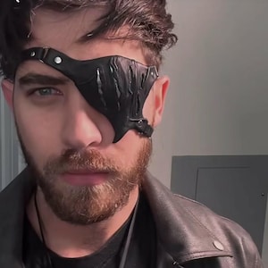 Può includere: Un uomo con la barba che indossa una giacca di pelle nera e una benda per l'occhio in pelle nera con un design decorativo.