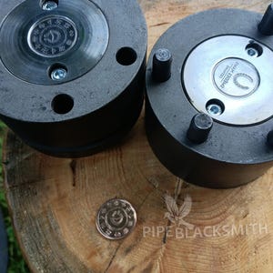 May include: Close-up of two large, dark gray metal components with circular designs and bolt fasteners. A small, bronze-colored coin with an intricate pattern rests nearby. The image is set on a wooden surface, with the text "PIPEBLACKSMITH" visible.